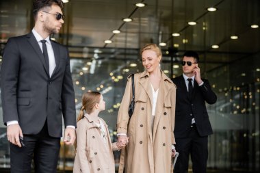 two bodyguards protecting successful woman and preteen kid, private security, happy blonde mother and daughter in trench coats near hotel, safety and protection, family travel, rich life 