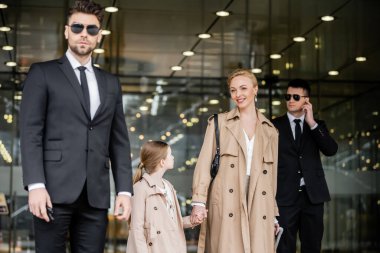 İki koruma başarılı kadın ve çocuğu koruyor, özel güvenlik, otelin yakınındaki trençkotlarda mutlu sarışın anne ve kız, güvenlik ve koruma, aile seyahati, zengin hayat 
