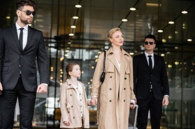 Başarılı bir kadın ve çocuğun yanında yürüyen iki koruma, otele giriş, özel güvenlik, trençkotlu sarışın anne ve kız, güvenlik ve koruma, aile seyahati, zengin hayat 