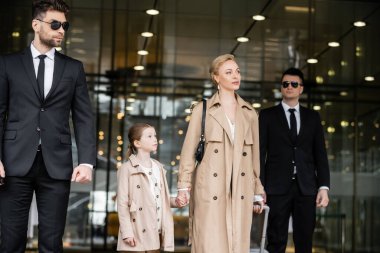 two bodyguards walking next to successful woman and preteen kid, entering hotel, private security, blonde mother and daughter in trench coats, safety and protection, family travel, rich life 