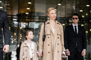 bodyguards walking next to blonde woman and preteen kid, entering hotel, private security, successful mother holding hands with daughter and wearing trench coats, safety and protection  clipart