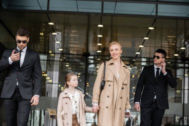 Korumalar, neşeli kadın ve çocuğun yanında yürürken, otele giriyorlar, özel güvenlik, anne kızıyla el ele tutuşuyor ve trençkotlar, emniyet ve koruma giyiyorlar. 