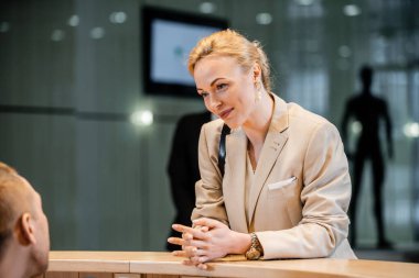 polite businesswoman talking to receptionist in hotel, hospitality industry, blonde and cheerful woman communicating with hotel staff, personal security, private safety, bodyguards on background  clipart