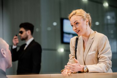 blonde businesswoman talking to receptionist in hotel, hospitality industry, attractive woman communicating with hotel staff, private security, personal safety, bodyguard on background  clipart