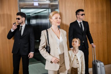 successful mother and child holding hands while standing near hotel elevator and bodyguards in suits, personal protection, woman and preteen daughter, family travel, security service  clipart