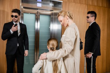 happy mother hugging child while standing near hotel elevator and bodyguards in suits, personal protection, successful woman and preteen daughter, family travel, security service  clipart