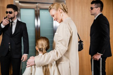 happy mother hugging child while standing near hotel elevator and bodyguards in suits, personal safety, successful woman and preteen daughter, family travel, security service 