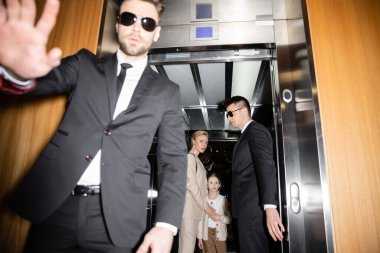 private security concept, handsome bodyguard in sunglasses and suit showing stop, no gesture to camera, protecting private space of famous clients in elevator of hotel, blurred mother and daughter  clipart