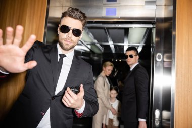 personal safety, handsome bodyguard in sunglasses and suit showing stop, no gesture to camera, protecting private space of famous clients in elevator of luxury hotel, blurred mother and daughter  clipart