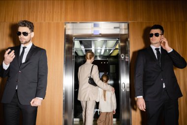 private safety, mother and daughter entering elevator of luxurious hotel, two bodyguards protecting their safety, handsome men in suits and sunglasses working in personal security service  clipart
