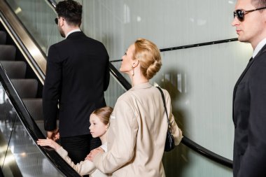 personal safety, blonde and successful woman and preteen girl standing on escalator of luxurious hotel, two bodyguards protecting safety of clients, rich lifestyle, conceptual, professionals  clipart