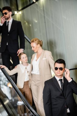 Kişisel güvenlik kavramı, kadın müşterilerin güvenliğini korurken iletişim kuran yakışıklı korumalar, zengin yaşam tarzı, sarışın ve başarılı kadın ve otelin yürüyen merdivenlerinde duran küçük kız.