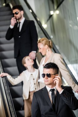 personal security concept, handsome bodyguards communicating while protecting safety of female clients, rich lifestyle, successful woman and preteen child standing on escalator of hotel