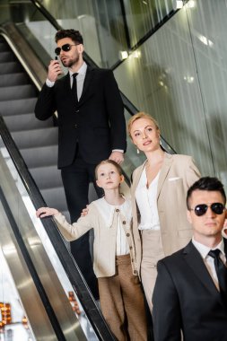 Kişisel güvenlik kavramı, başarılı kadın ve küçük kızı lüks otelin yürüyen merdiveninde duruyorlar, iki koruma müşterilerin güvenliğini sağlarken iletişim kuruyorlar, zengin yaşam tarzı. 