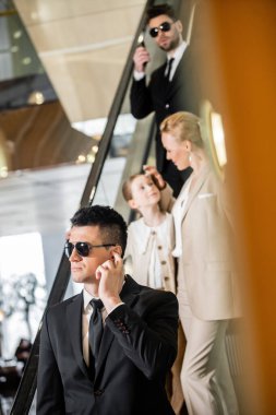 personal security concept, bodyguards communicating while protecting safety of clients, rich lifestyle, blonde and successful woman and preteen daughter standing on escalator of luxurious hotel  clipart