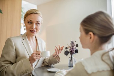 Anne ve kızı birlikte kaliteli zaman geçiriyorlar, sarışın kadın ergen kızla konuşuyor ve elinde bir fincan kahve, çalışan ebeveyn ve çocuk, modern ebeveynlik, aile bağı 