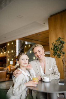 Anne ve kızı birlikte kaliteli zaman geçiriyor, sarışın kadın reşit olmayan kıza sarılıyor ve içki ve akıllı telefonun yanındaki kameraya bakıyor, çalışan ebeveyn ve çocuk, modern ebeveynlik, aile bağı 