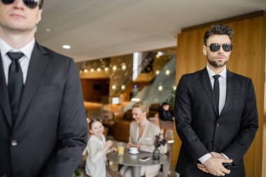 Güvenlik hizmeti, özel güvenlik konsepti, resmi giysili yakışıklı korumalar ve güneş gözlükleri bulanık arka planda müşterilerin güvenliğini koruyor, anne ve kızı kafede. 