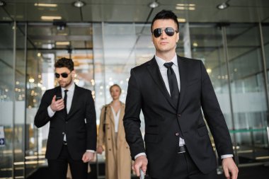 private security service, personal safety concept, handsome bodyguards in formal wear and sunglasses protecting safety of client on blurred background, walking in hotel  clipart