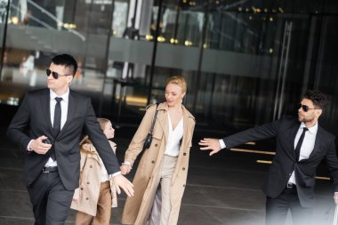 handsome bodyguards protecting safety of female clients, blonde woman and daughter walking out of hotel, safety of rich family, men in sunglasses and suits on duty, work in security  clipart