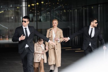 Özel güvenlik, kadın müşterileri koruyan kişisel korumalar, otelden çıkan sarışın kadın ve kız, zengin ailenin güvenliği, resmi giyinen erkekler ve işini yapan güneş gözlükleri. 
