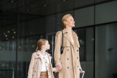 modern fashion, mother daughter time, blonde woman with luggage holding hand of preteen girl while walking out of hotel together, smart casual, beige trench coats, outerwear, modern parenting  clipart