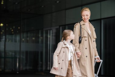 autumn fashion, mother daughter time, positive woman with luggage holding hand of preteen girl while walking out of hotel together, smart casual, beige trench coats, outerwear, modern parenting  clipart