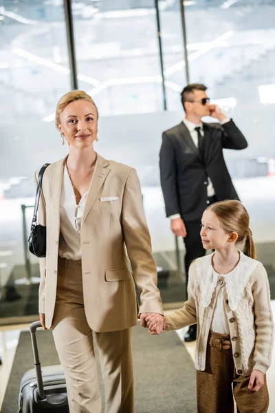 luxury lifestyle, rich life, personal security, cheerful woman with child, blonde woman holding hands with girl and walking inside of hotel, bodyguard in suit standing on blurred background