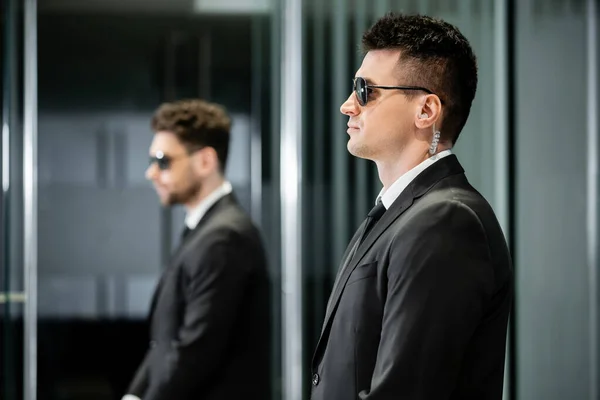 bodyguard service, private security, professional in suit and sunglasses standing in hotel lobby near work partner, earpiece, communication, luxury hotel, vigilance, protection and work, side view