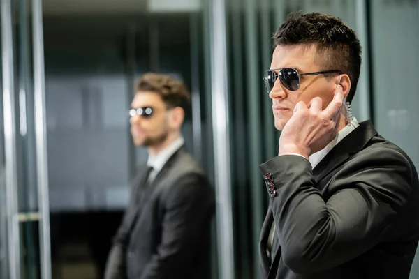 bodyguard service, private security, professional guards in suits and sunglasses standing in hotel lobby, handsome man with earpiece communicating with work partner, luxury hotel, vigilance