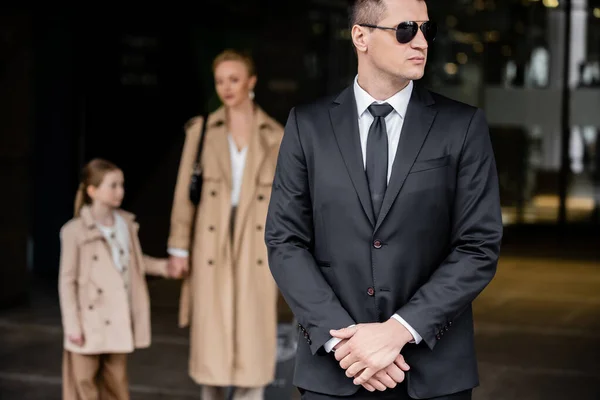 personal security service, lifestyle, bodyguard in suit standing near successful woman with preteen child, protecting mother and daughter near hotel, rich life, family travel, private security