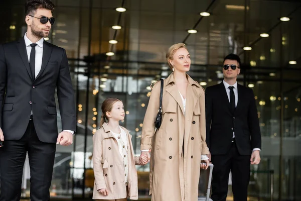 stock image two bodyguards walking next to successful woman and preteen kid, entering hotel, private security, blonde mother and daughter in trench coats, safety and protection, family travel, rich life 
