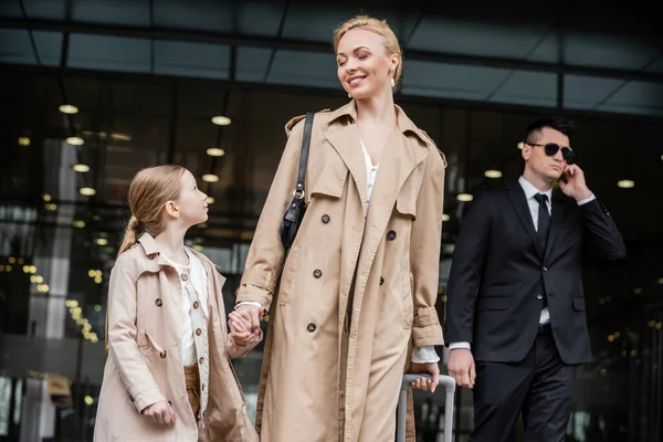 Stock image personal security, lifestyle, blonde mother with preteen girl holding hands near hotel, successful woman and child, bodyguard in suit standing on blurred background, rich life 