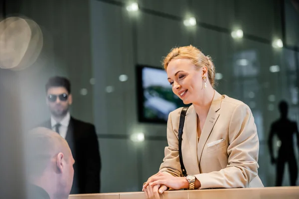 cheerful businesswoman talking to receptionist in hotel, hospitality industry, blonde and polite woman communicating with hotel staff, personal security, private safety, bodyguards on background