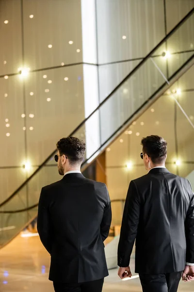security management of luxury hotel, back view of two men in formal wear and sunglasses, bodyguards on duty, safety measures, vigilance, black suits, private security, strong guards