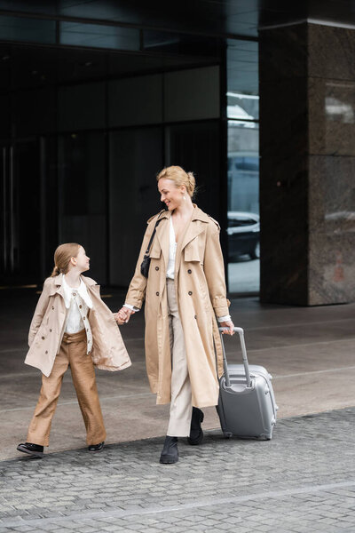 trendy look, mother daughter time, happy woman with luggage holding hand of preteen girl while walking out of hotel together, smart casual, beige trench coats, outerwear, modern parenting 