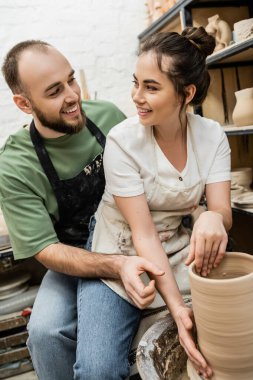 Smiling craftspeople looking at each other and making clay vase on pottery wheel in ceramic studio clipart