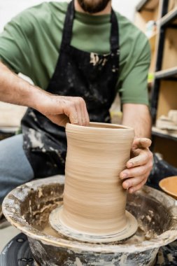Partial view of blurred artisan in apron shaping clay on pottery wheel in ceramic workshop clipart