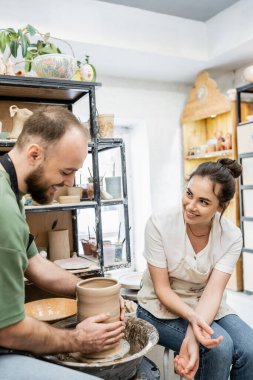 Joyful artisan in apron talking to boyfriend making clay vase on pottery wheel in ceramic workshop clipart