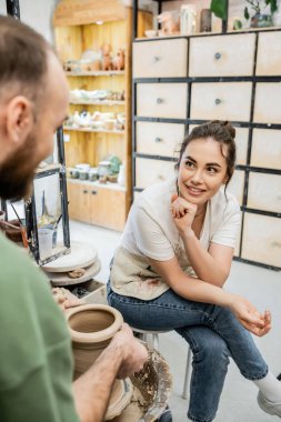 Positive craftsman in apron talking to girlfriend making clay vase on pottery wheel in workshop clipart