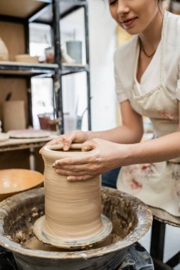 Cropped view of female potter in apron creating clay vase on pottery wheel in blurred workshop clipart