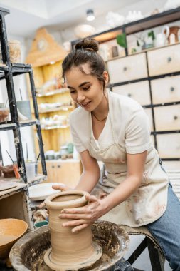 Positive female artisan in apron shaping clay vase on pottery wheel in ceramic studio at background clipart