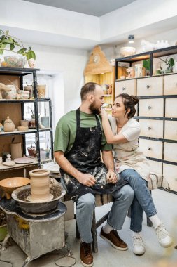 Craftswoman in apron hugging boyfriend and talking near clay on pottery wheel in workshop clipart