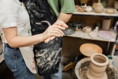 Cropped view of couple of sculptors in aprons holding hands near blurred  pottery wheel in workshop clipart