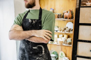 Cropped view of bearded artisan in apron crossing arms while standing in ceramic workshop clipart