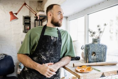 Sakallı zanaatkar seramik atölyesinde elinde çiçeklerle vazonun yanında dikilirken gözünü kaçırıyor.