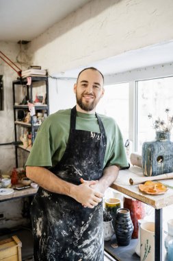 Smiling craftsman in apron looking at camera and standing near vases and window in ceramic workshop clipart