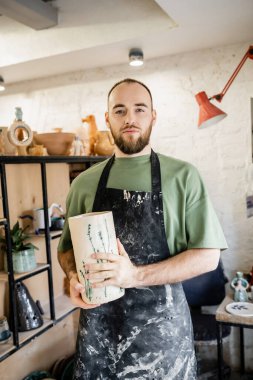 Bearded craftsman in apron holding ceramic vase while standing and looking at camera in workshop clipart