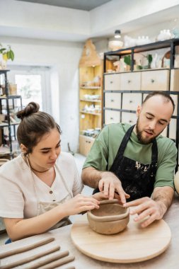 Couple of potters in aprons shaping clay bowl together while working in ceramic workshop clipart