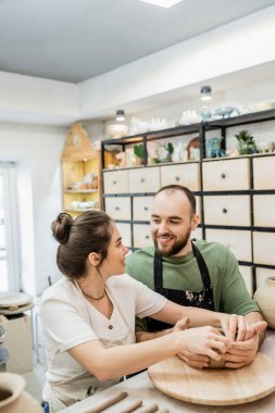 Smiling couple of artisans in aprons making clay bowl and looking at each other in ceramic studio clipart